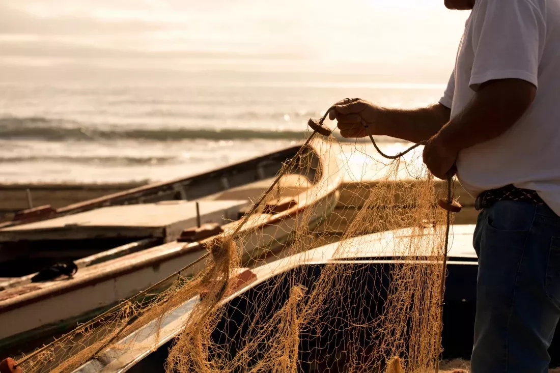 Pescador desenreda una red de pesca junto a barcas en la playa al atardecer